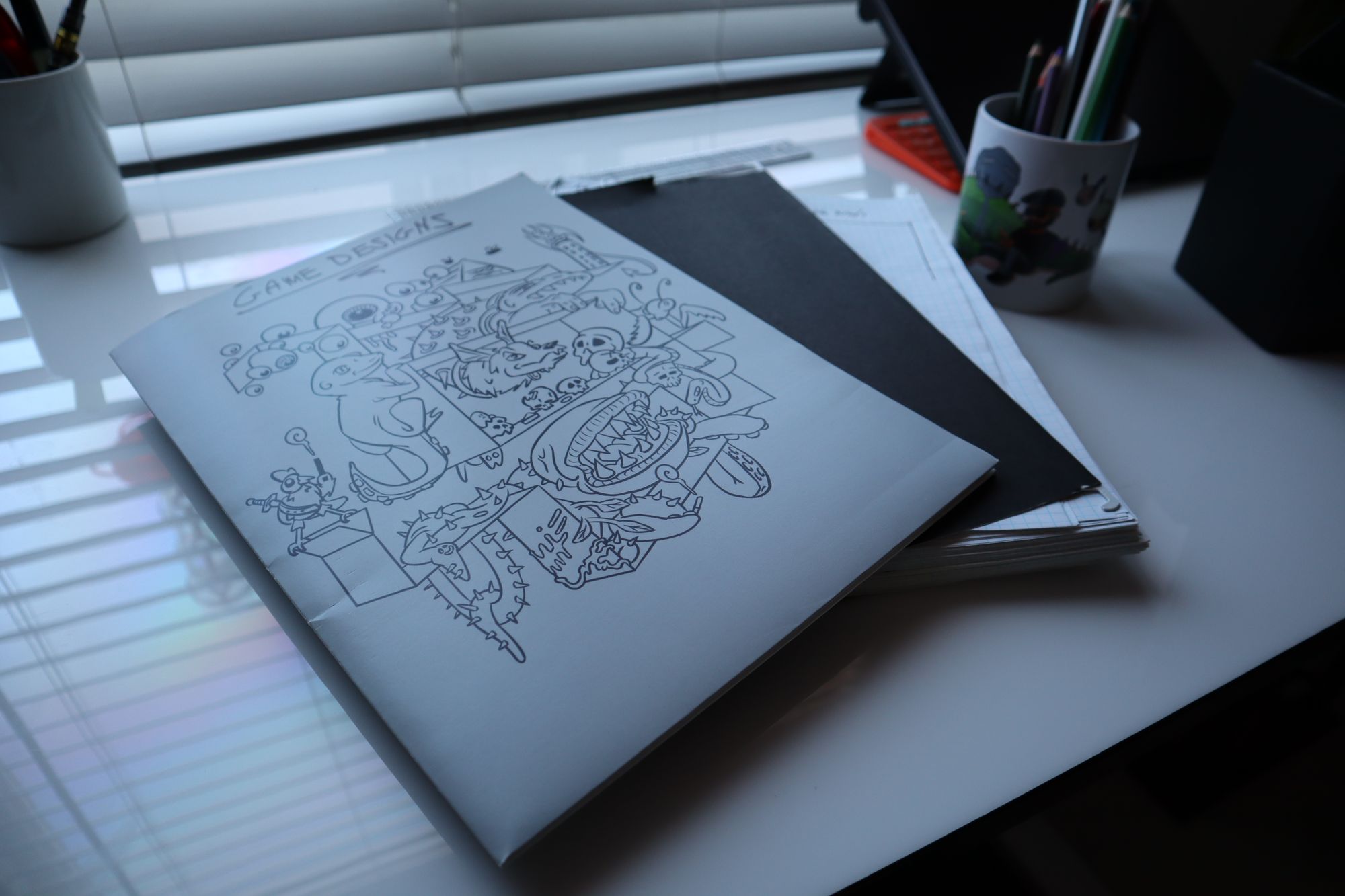A photo of a desk with some clutter like mugs with pencils and an iPad in the corner. In the center is a stack of graph paper, and a folder with some doodles on the cover, labeled "GAME DESIGNS".